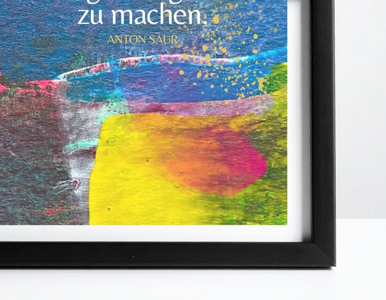 Schwarzer Bilderrahmen mit einem farbenfrohen, abstrakten Kunstwerk, das hauptsächlich blaue, gelbe, pinke und grüne Farbtöne zeigt, mit einem Teil eines Zitats von Anton Saur.